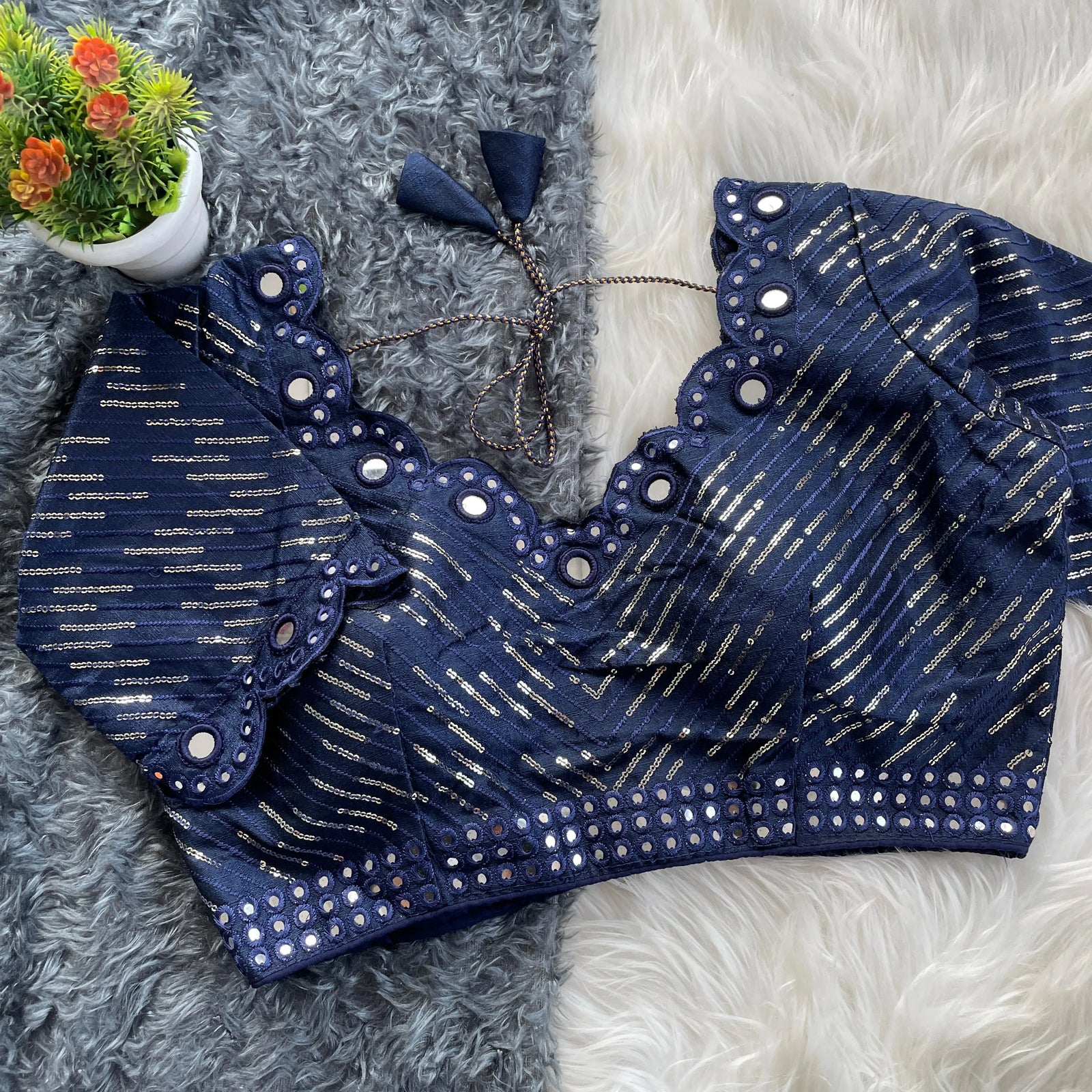 Navy blue blouse with silver sequin stripes, scalloped neckline embellished with mirror work, decorative sleeve borders, and tie-up tassels, displayed on a textured grey and white fur background with a small potted plant nearby.