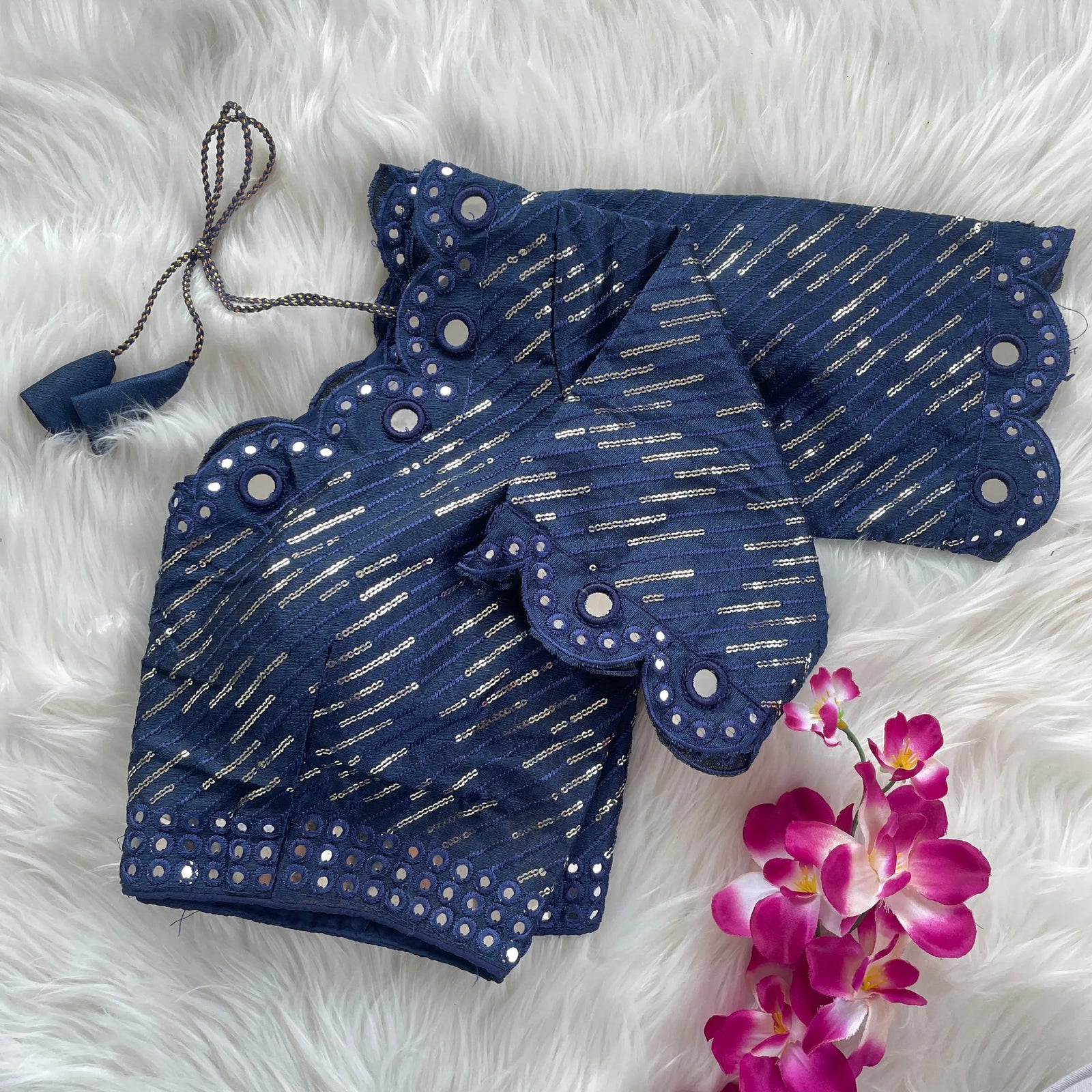 Navy blue blouse with silver sequin stripes, scalloped neckline embellished with mirror work, decorative sleeve borders, and tie-up tassels, displayed on a textured grey and white fur background with a small potted plant nearby

