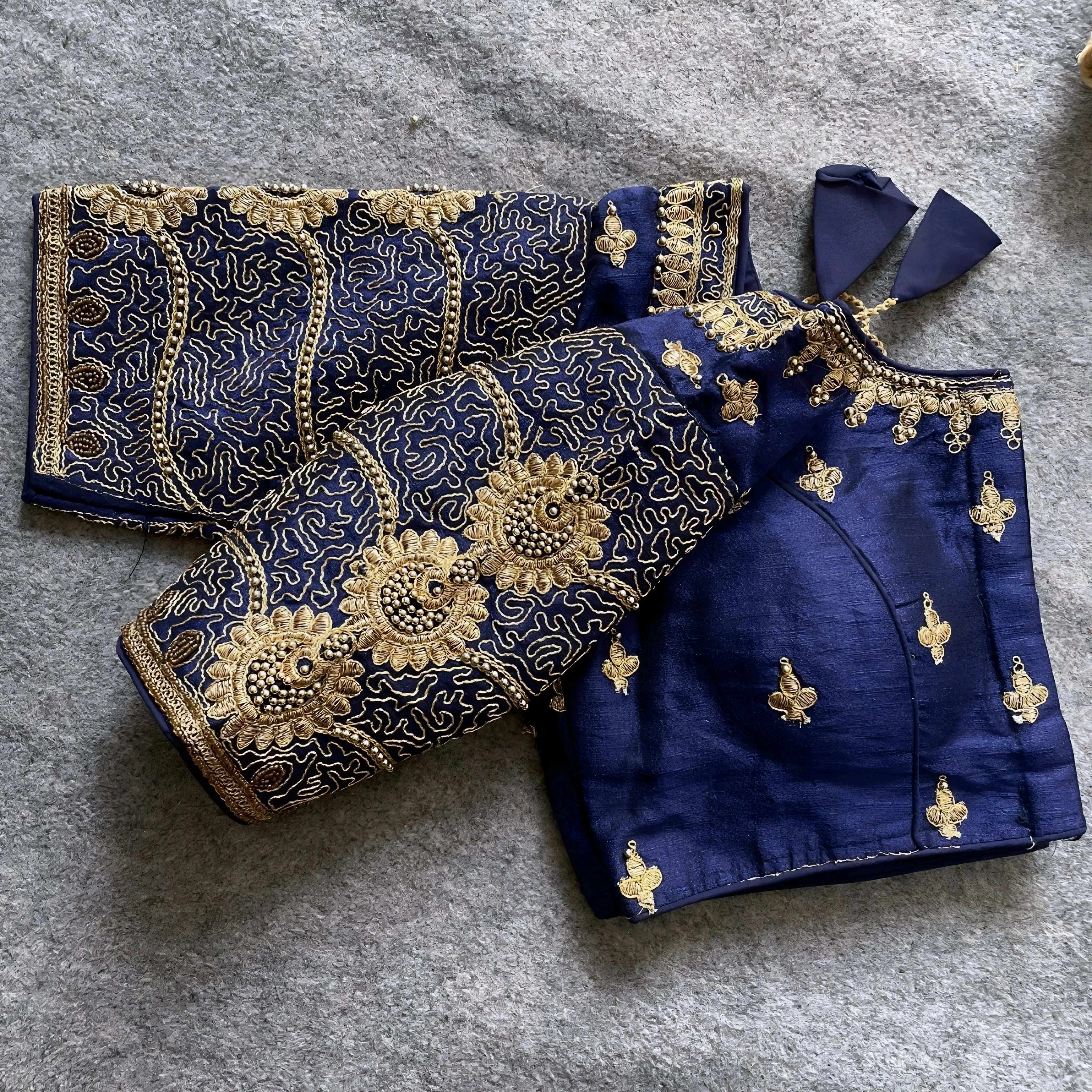 Traditional navy blue silk blouse featuring intricate gold embroidery and beadwork with floral and paisley motifs on the sleeves and neckline, displayed flat on a textured surface with a bunch of artificial flowers and a woven mat in the background.