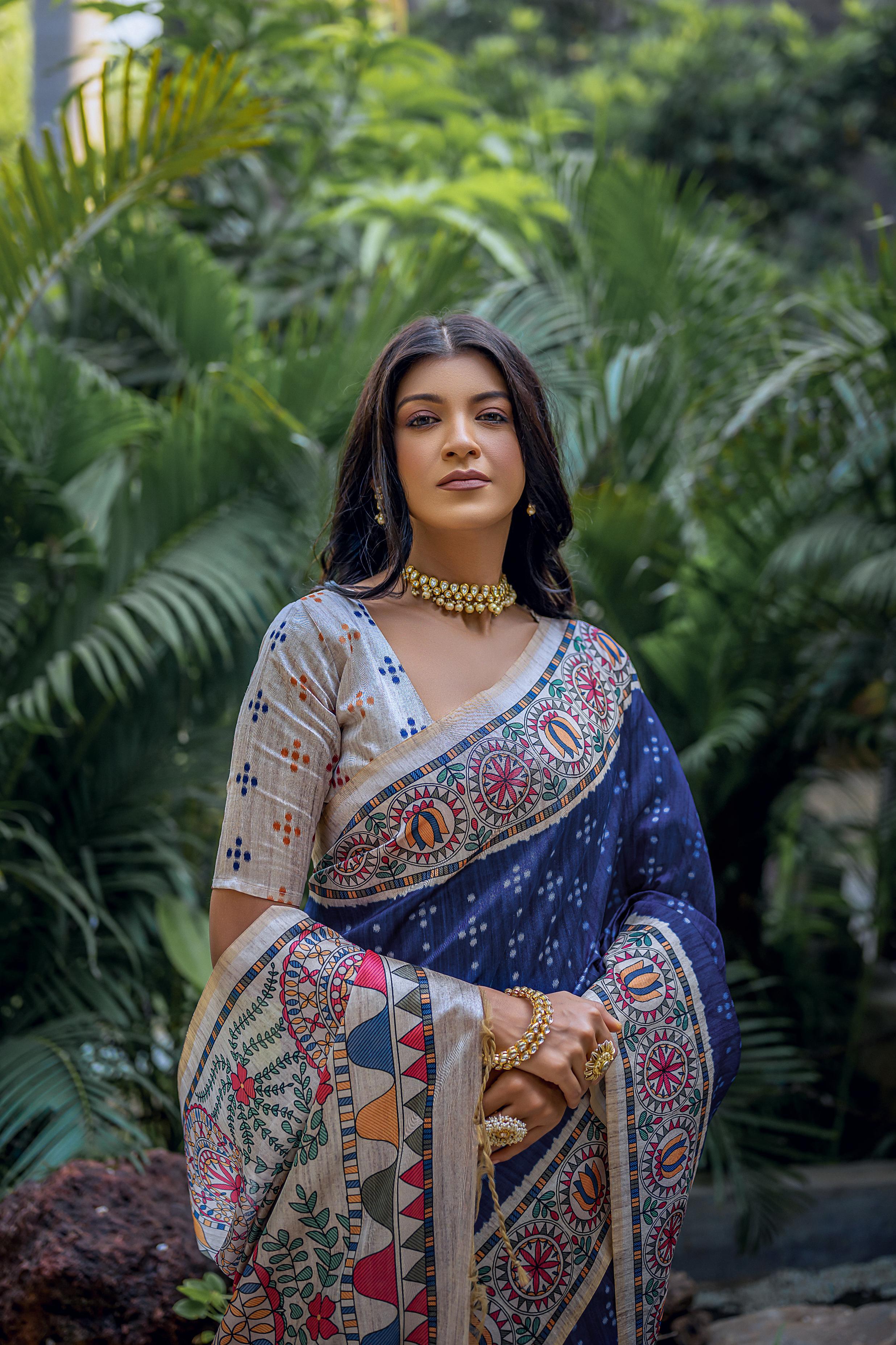 A woman models a deep Navy Blue Tussar Silk Saree with small scattered prints. The pallu features a wide, elaborate panel with intricate Madhubani-style folk art prints in white, red, green, and blue. She wears a cream/gold embellished blouse and a gold Kundan choker necklace.