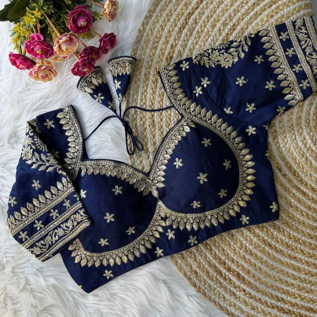 Navy blue blouse with intricate gold zari embroidery featuring floral and leaf motifs, decorative borders, and tie-up tassels, displayed on a textured mat with pink flowers in the background.