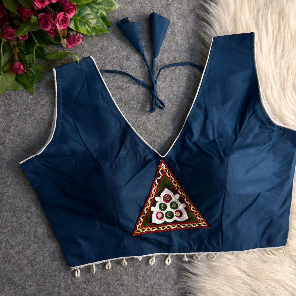  Navy blue padded blouse with white piping, featuring a central embroidered triangle motif with mirror work and shell detailing along the hem, displayed on a grey surface with artificial pink flowers and white fur in the background.