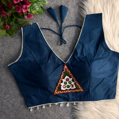  Navy blue padded blouse with white piping, featuring a central embroidered triangle motif with mirror work and shell detailing along the hem, displayed on a grey surface with artificial pink flowers and white fur in the background.