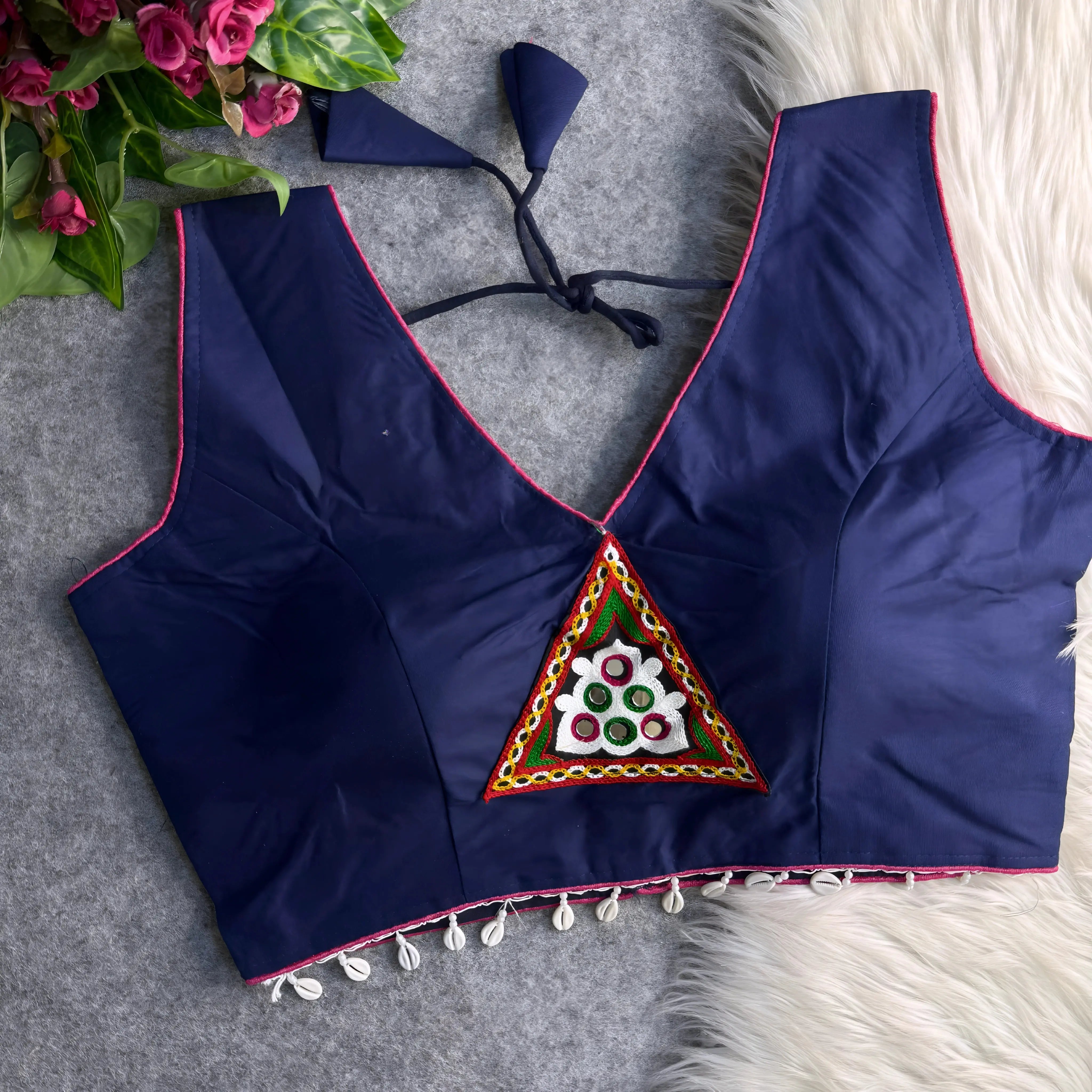 Navy blue padded blouse with pink piping, featuring a central embroidered triangle motif with mirror work and shell detailing along the hem, displayed on a grey surface with artificial pink flowers and white fur in the background.