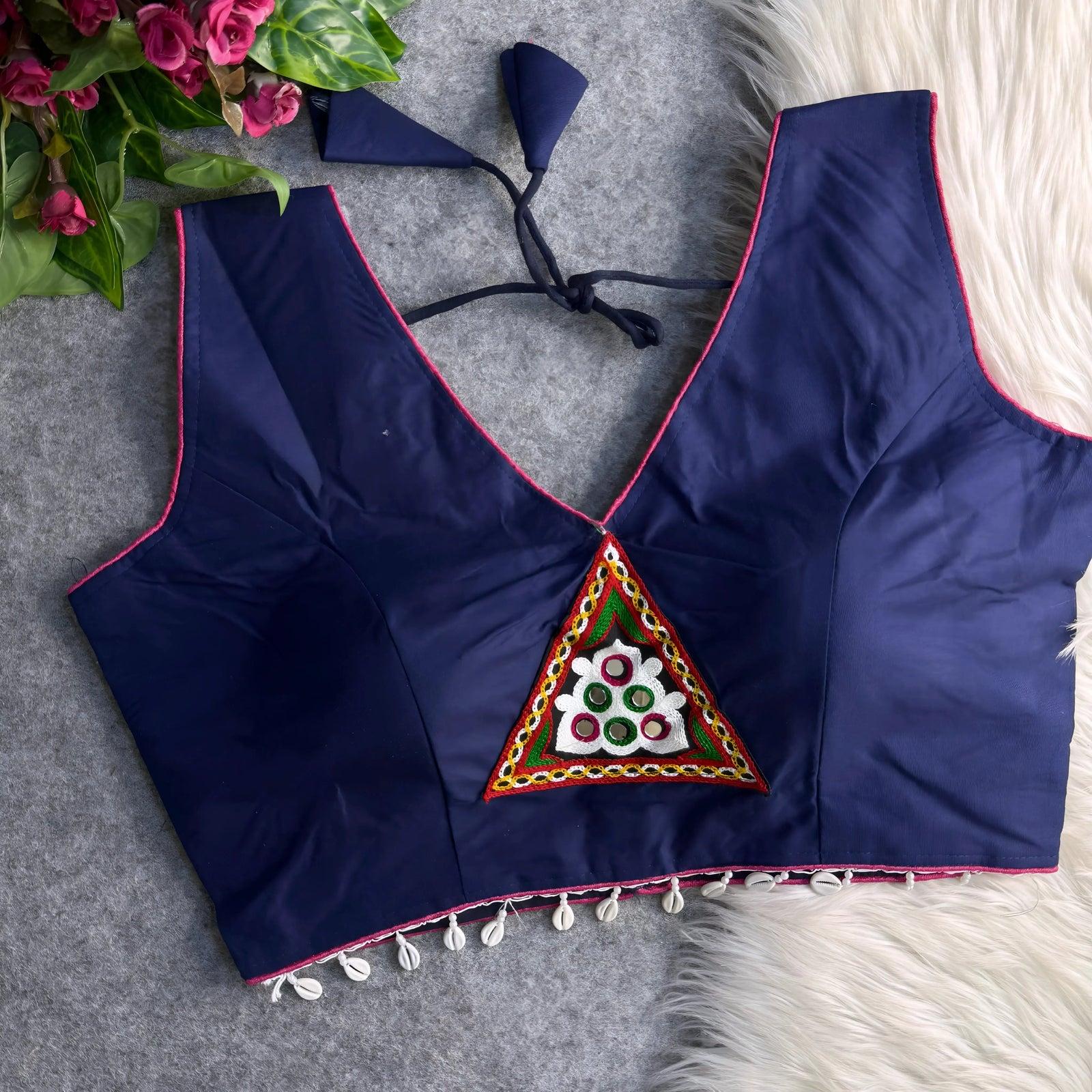 Navy blue padded blouse with pink piping, featuring a central embroidered triangle motif with mirror work and shell detailing along the hem, displayed on a grey surface with artificial pink flowers and white fur in the background.