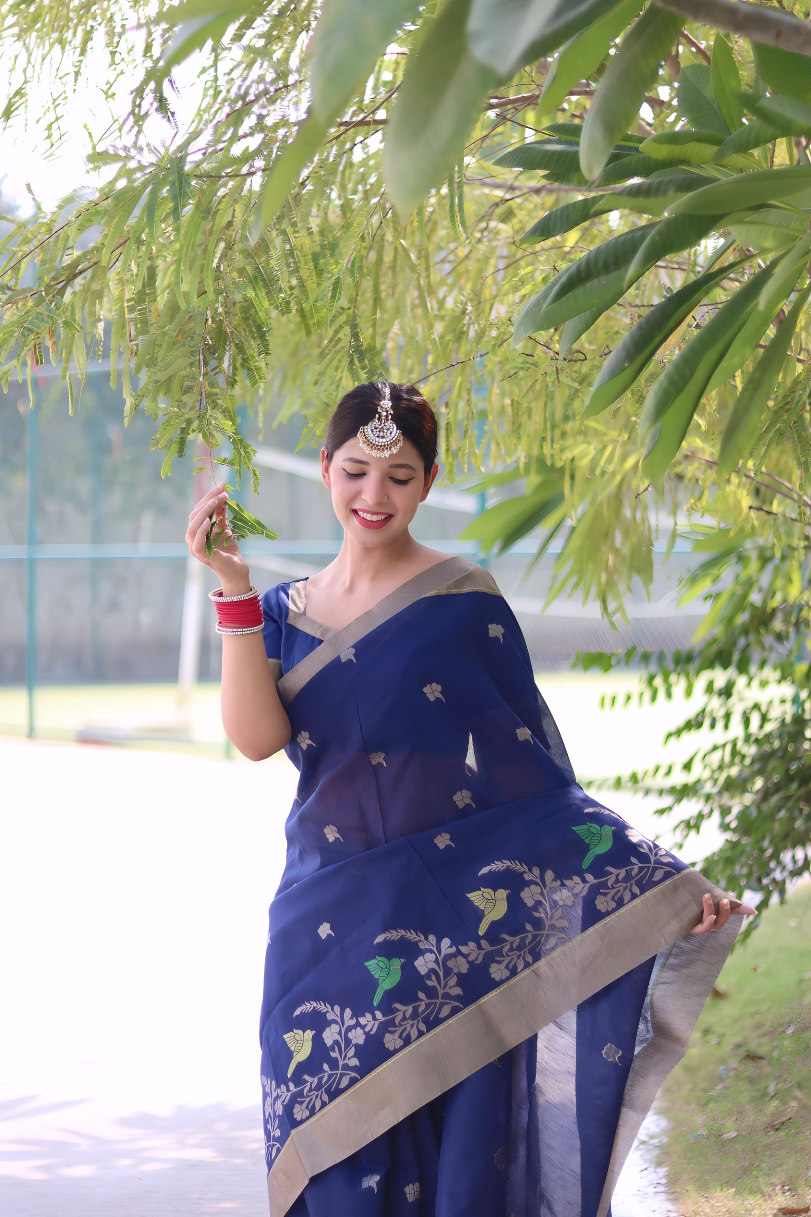 A woman models a vibrant Navy Blue Linen Silk Saree with delicate gold/silver Zari woven motifs and a wide metallic silver border. She wears a beige blouse, red and white bangles, and a traditional headpiece, posing outdoors under a tree.