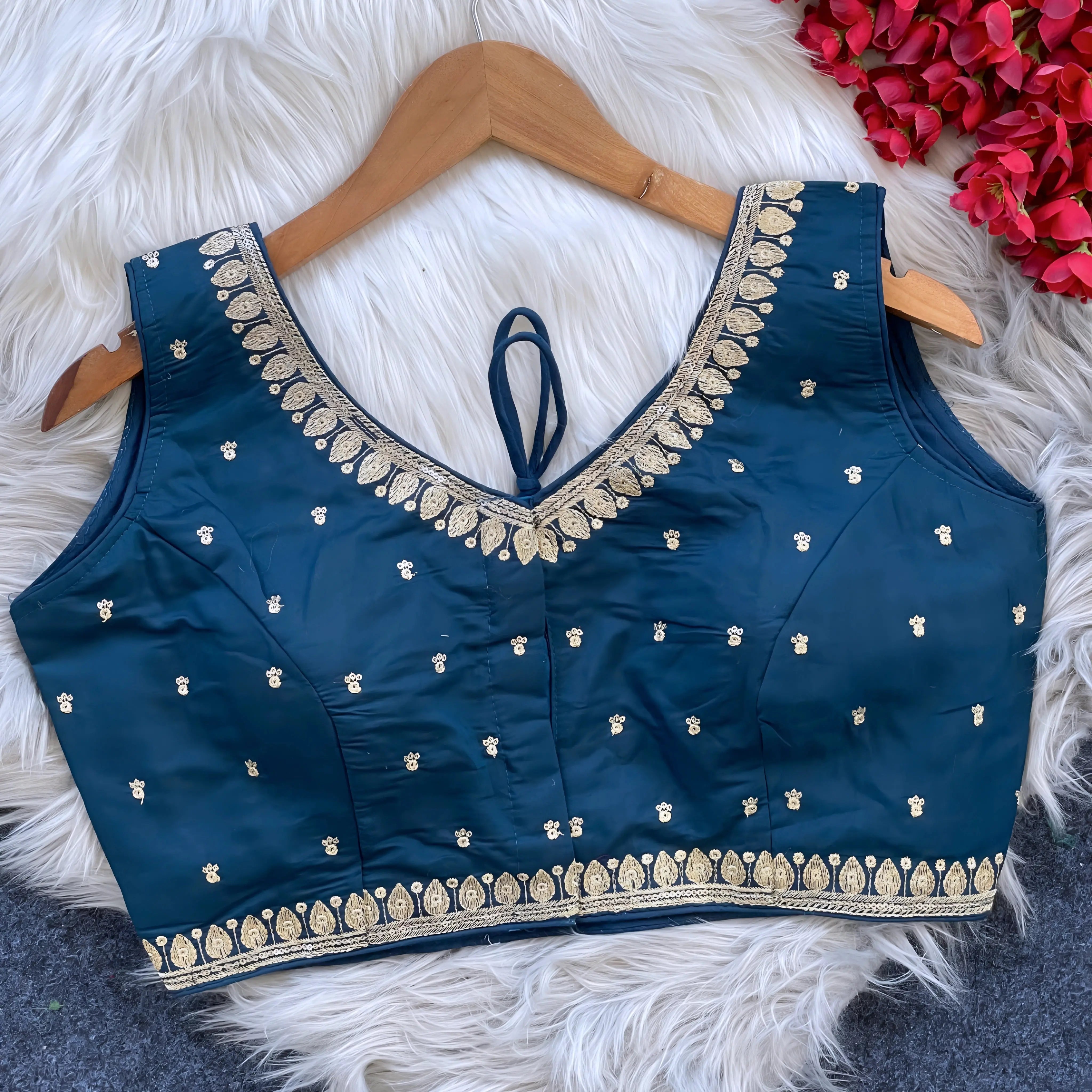 Navy blue padded blouse with intricate gold embroidery along the neckline and hem, featuring small gold embroidered motifs, displayed on a wooden hanger over a white fur surface with red artificial flowers in the background.