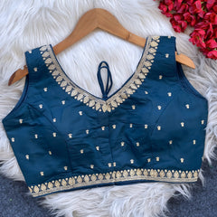 Navy blue padded blouse with intricate gold embroidery along the neckline and hem, featuring small gold embroidered motifs, displayed on a wooden hanger over a white fur surface with red artificial flowers in the background.