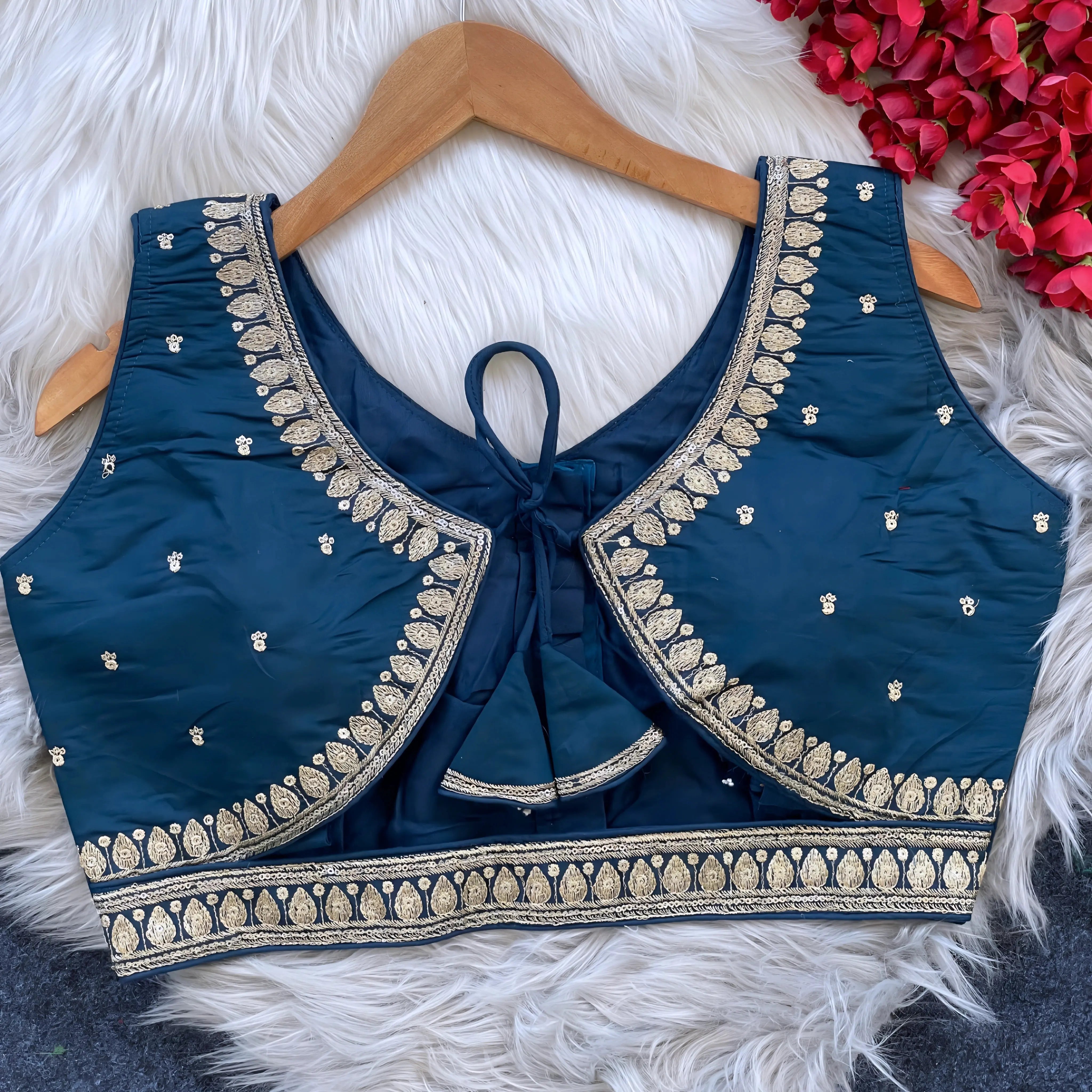 Navy blue padded blouse with intricate gold embroidery along the neckline and hem, featuring small gold embroidered motifs, displayed on a wooden hanger over a white fur surface with red artificial flowers in the background.
