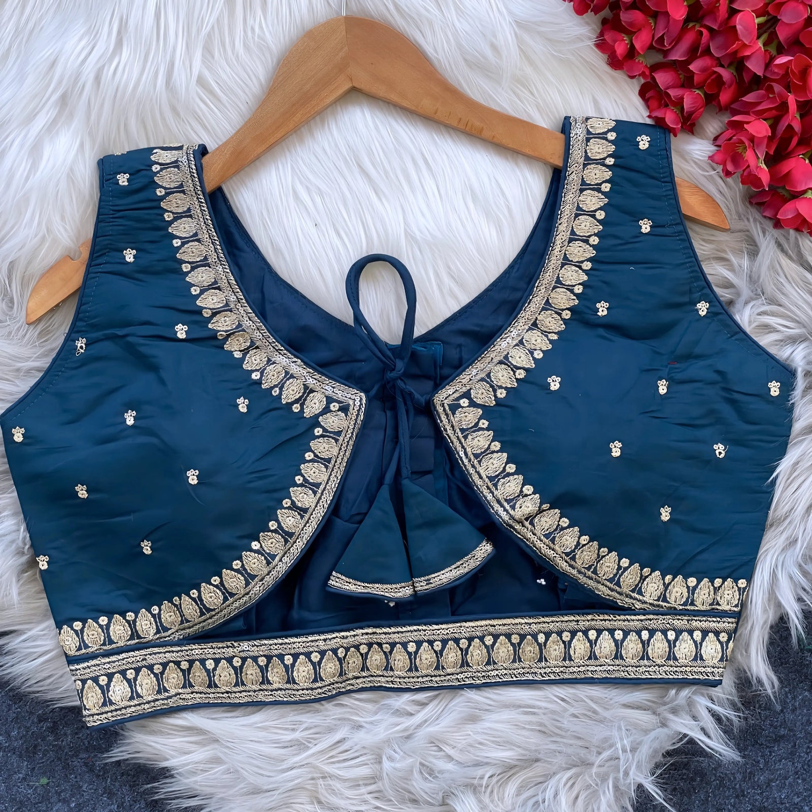 Navy blue padded blouse with intricate gold embroidery along the neckline and hem, featuring small gold embroidered motifs, displayed on a wooden hanger over a white fur surface with red artificial flowers in the background.