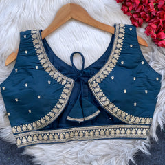 Navy blue padded blouse with intricate gold embroidery along the neckline and hem, featuring small gold embroidered motifs, displayed on a wooden hanger over a white fur surface with red artificial flowers in the background.