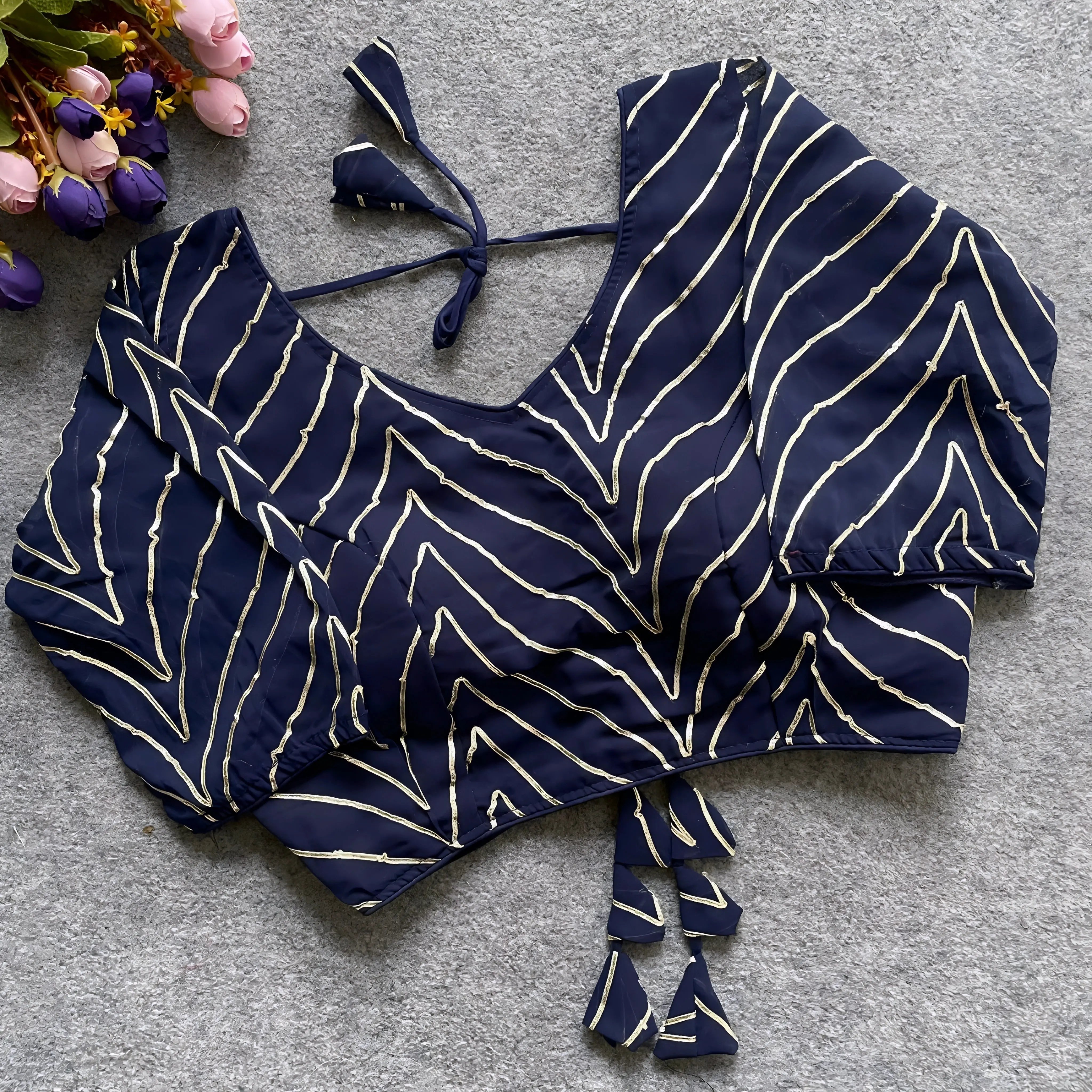 Navy blouse beautifully adorned with gold gota patti zigzag embroidery, three-quarter sleeves, and a stylish back tie-up with matching triangular tassels. Displayed flat on a grey textured surface beside pink and purple flowers, showcasing its festive and sophisticated ethnic design.