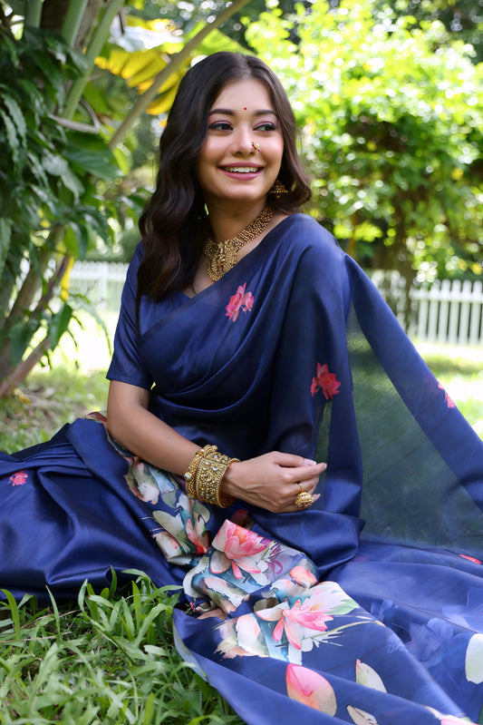 A woman models a stunning Deep Navy Blue Soft Tussar Silk Saree. The saree features a large-scale digital print of colorful floral and lotus motifs in pink, white, and green on the pallu and lower drape. She wears a matching blue blouse and heavy gold Kundan jewelry, posing outdoors in a garden setting.