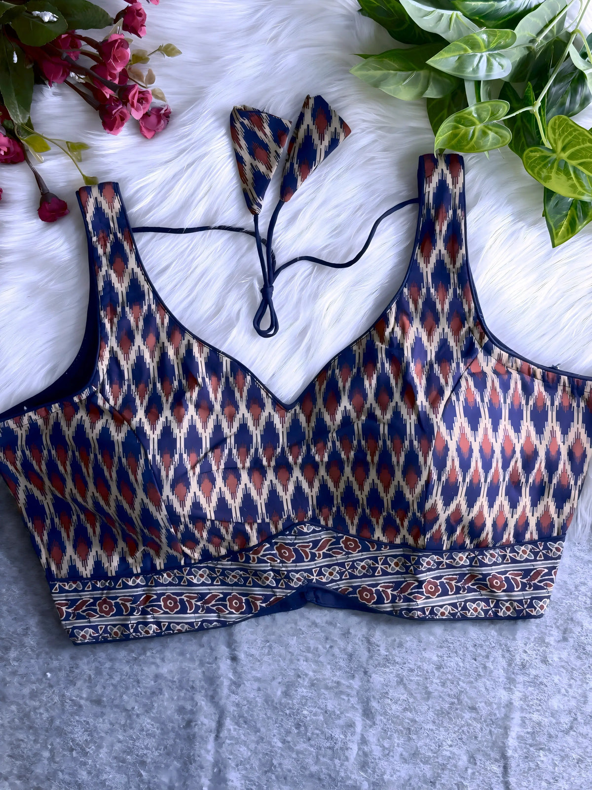 Navy blue silk blouse featuring a traditional Ikat-inspired geometric print in cream, maroon, and gold, with wide shoulder straps and contoured cups. The design has a deep neckline, a contrasting floral patterned border at the hem, and a matching triangular tasseled tie-back, displayed flat on a white fur background with artificial pink flowers and green foliage arranged at the corners.