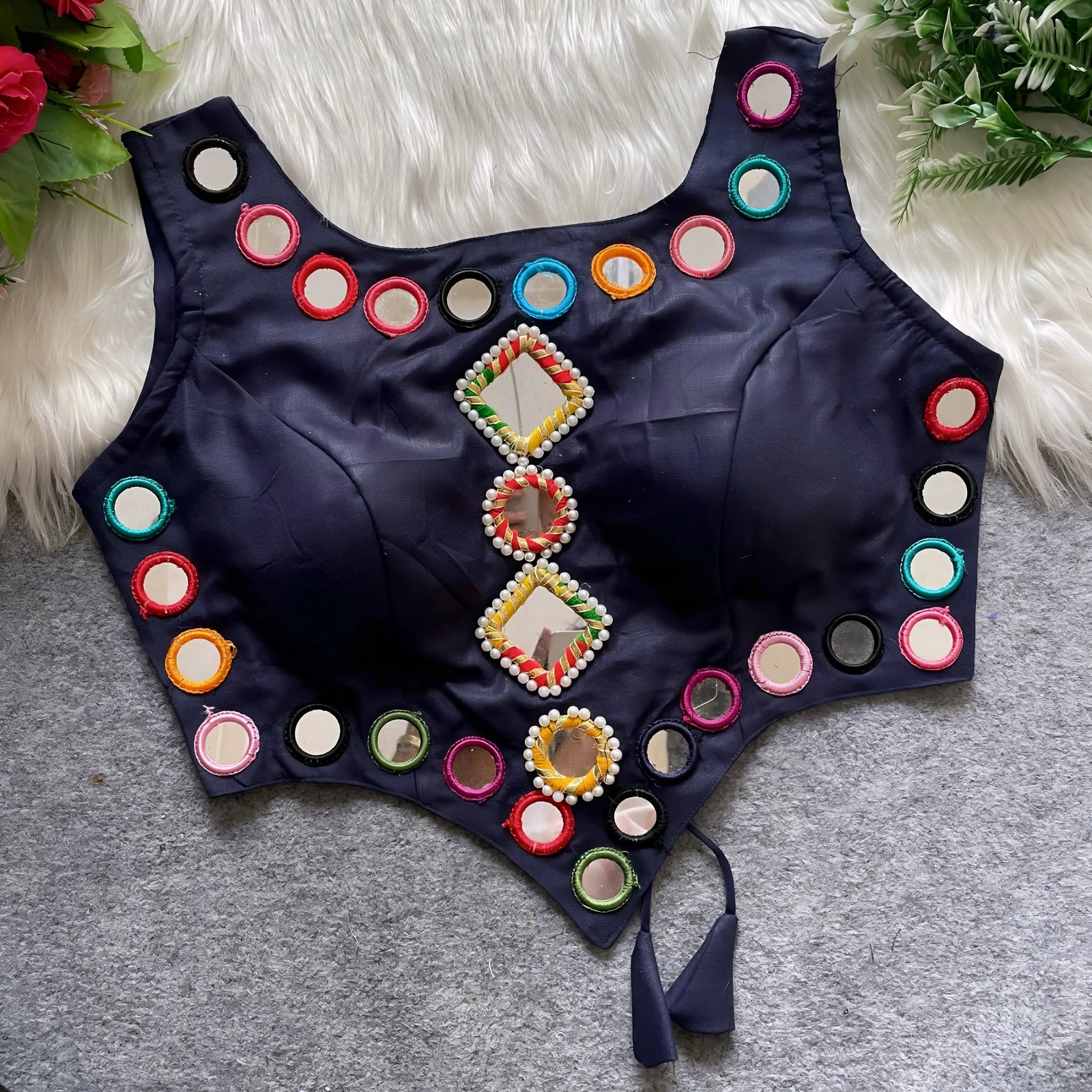 Navy blue ethnic blouse featuring intricate mirror work with multicolored embroidered borders and decorative diamond and round mirrors arranged vertically at the center, accented with pearl details and a tie-up tassel at the waist. Displayed flat on a grey surface with a white faux fur background and green leafy accents, highlighting its festive and traditional design.