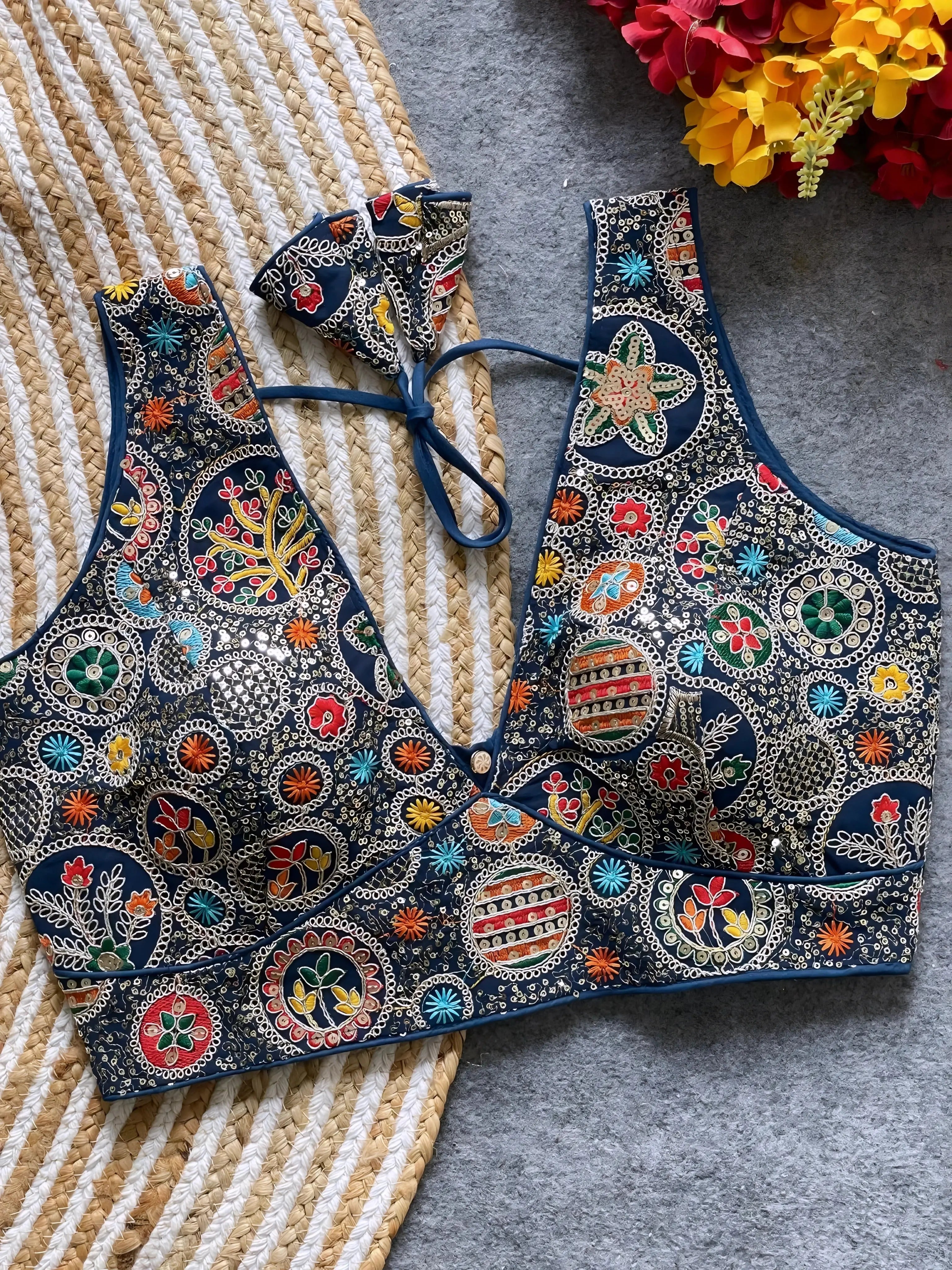 Traditional navy blue blouse featuring vibrant multicolor thread embroidery and mirror work in circular floral and geometric patterns, with a deep V-neck and tie-back detail, displayed flat on a textured mat with artificial red and yellow flowers in the corner.