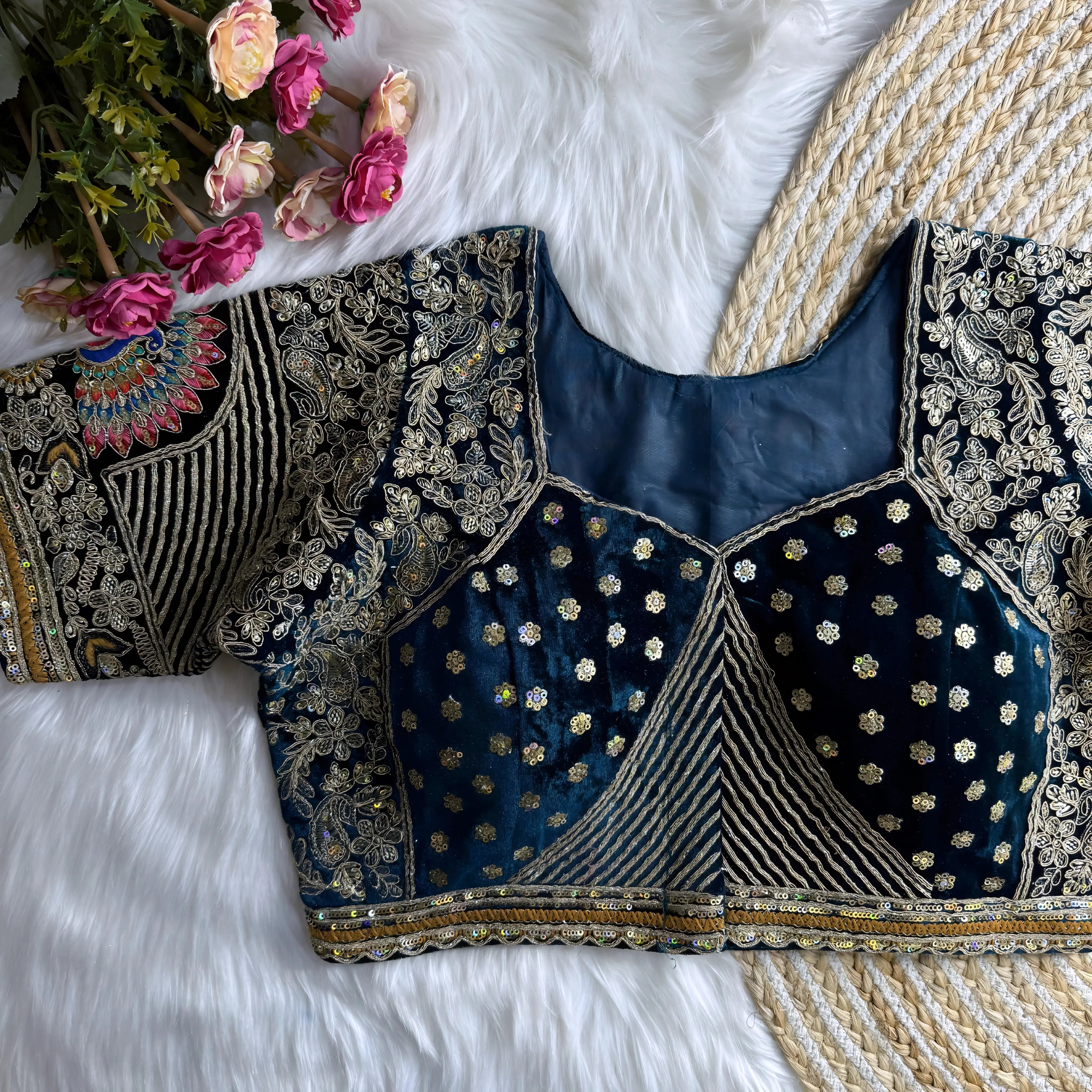 Traditional navy blue silk blouse featuring intricate gold zardosi embroidery with floral and leaf motifs, sequin embellishments, geometric panel designs, and a detailed multicolor peacock feather motif on the sleeve, displayed flat on a white fur surface with a braided mat and artificial pink flowers in the corner.