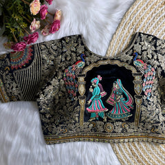 Traditional navy blue silk blouse featuring intricate gold zardosi embroidery with floral and leaf motifs, sequin embellishments, geometric panel designs, and a detailed multicolor peacock feather motif on the sleeve, displayed flat on a white fur surface with a braided mat and artificial pink flowers in the corner.