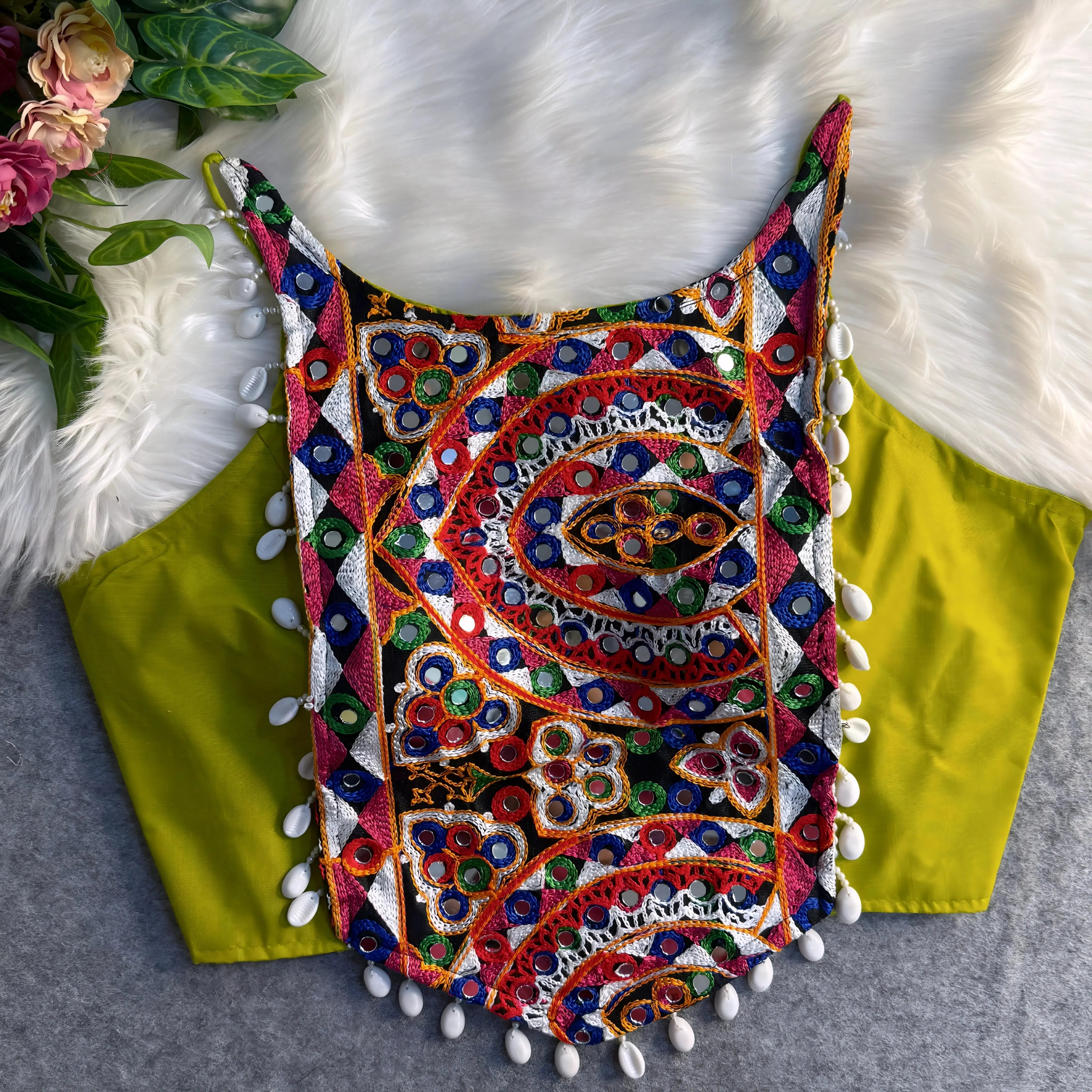 Traditional neon green blouse featuring vibrant multicolor mirror work and intricate thread embroidery in geometric and floral patterns, accented with white shell tassels along the hem, displayed flat on a soft fur and grey background with floral and leafy decor.