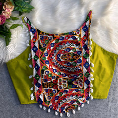 Traditional neon green blouse featuring vibrant multicolor mirror work and intricate thread embroidery in geometric and floral patterns, accented with white shell tassels along the hem, displayed flat on a soft fur and grey background with floral and leafy decor.