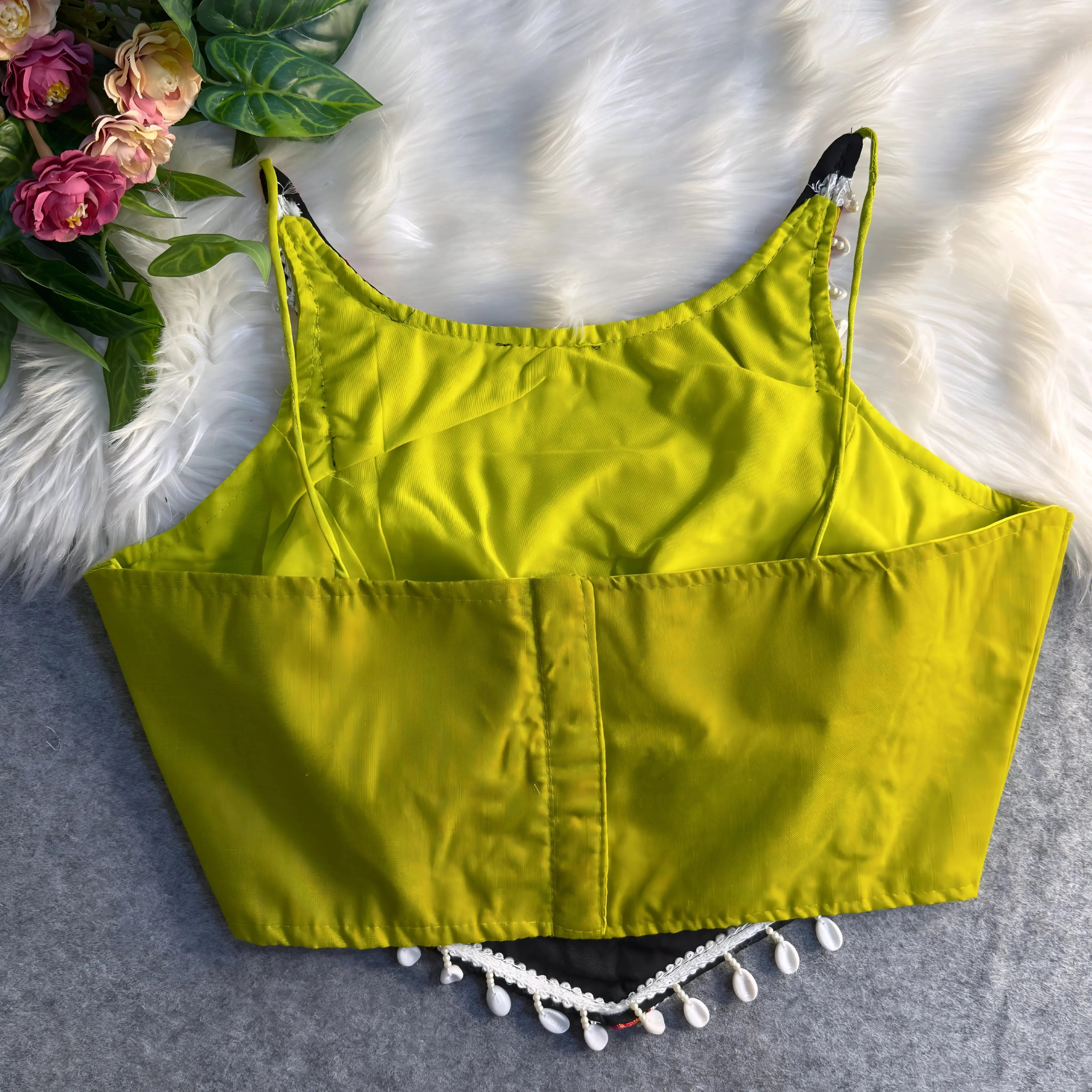 Traditional neon green blouse featuring vibrant multicolor mirror work and intricate thread embroidery in geometric and floral patterns, accented with white shell tassels along the hem, displayed flat on a soft fur and grey background with floral and leafy decor.