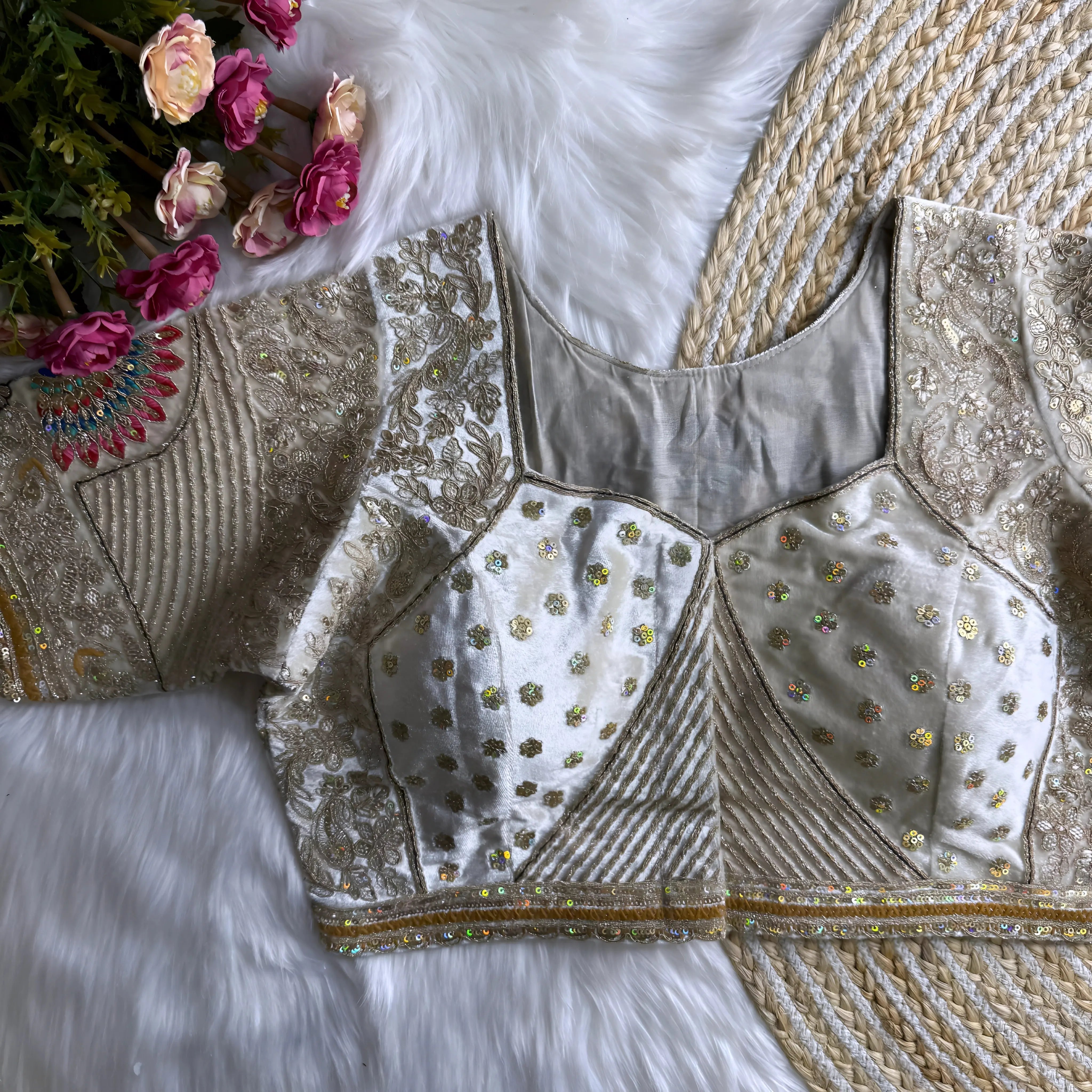 Traditional off-white blouse featuring intricate gold thread embroidery, sequin floral motifs, geometric panel designs, and subtle mirror work, displayed flat on a furry white surface with a braided natural mat and surrounded by pink artificial flowers in the corner.