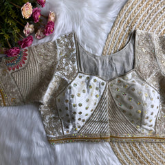 Traditional off-white blouse featuring intricate gold thread embroidery, sequin floral motifs, geometric panel designs, and subtle mirror work, displayed flat on a furry white surface with a braided natural mat and surrounded by pink artificial flowers in the corner.