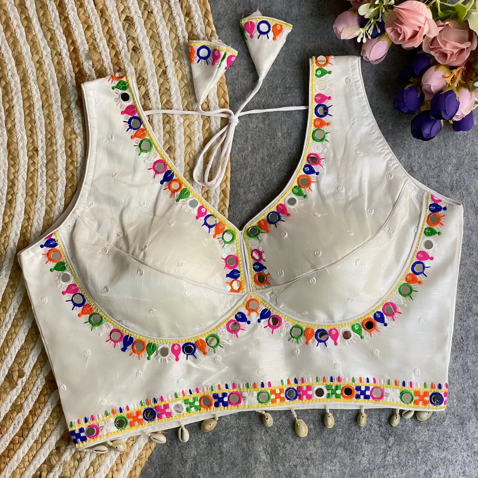 Off-white blouse featuring vibrant mirror work and traditional Kutch multicolor embroidery along the neckline and hem, with delicate sequin accents and shell tassels. The blouse includes a stylish back tie-up with matching embroidered tassels and is displayed flat on a textured surface with a floral bouquet, highlighting its festive ethnic elegance.