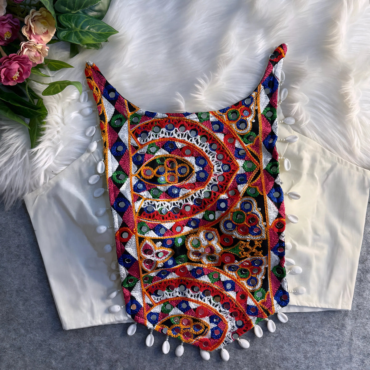 Traditional off-white blouse featuring vibrant multicolor mirror work and intricate thread embroidery in geometric and paisley patterns, accented with white shell tassels along the hem, displayed flat on a soft fur and grey background with floral and leafy decor.