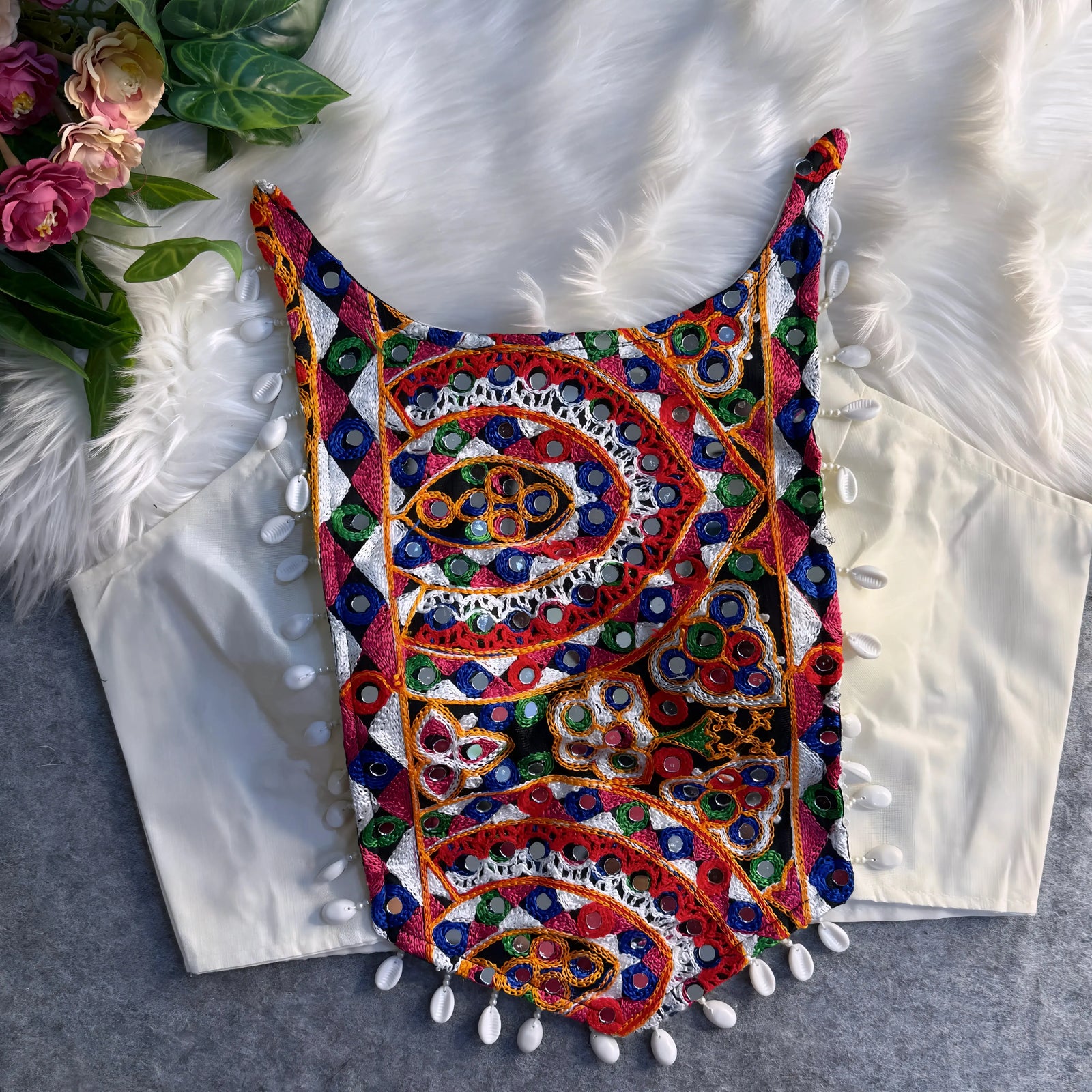 Traditional off-white blouse featuring vibrant multicolor mirror work and intricate thread embroidery in geometric and paisley patterns, accented with white shell tassels along the hem, displayed flat on a soft fur and grey background with floral and leafy decor.