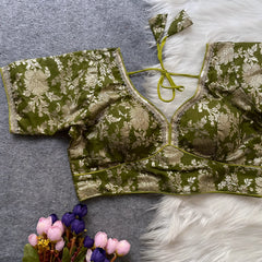 Olive green brocade blouse featuring intricate gold floral zari work, decorative trim along the neckline, and short sleeves displayed flat on a grey surface with a white faux fur accent and a bunch of artificial pink and purple flowers for an elegant, festive ethnic look.