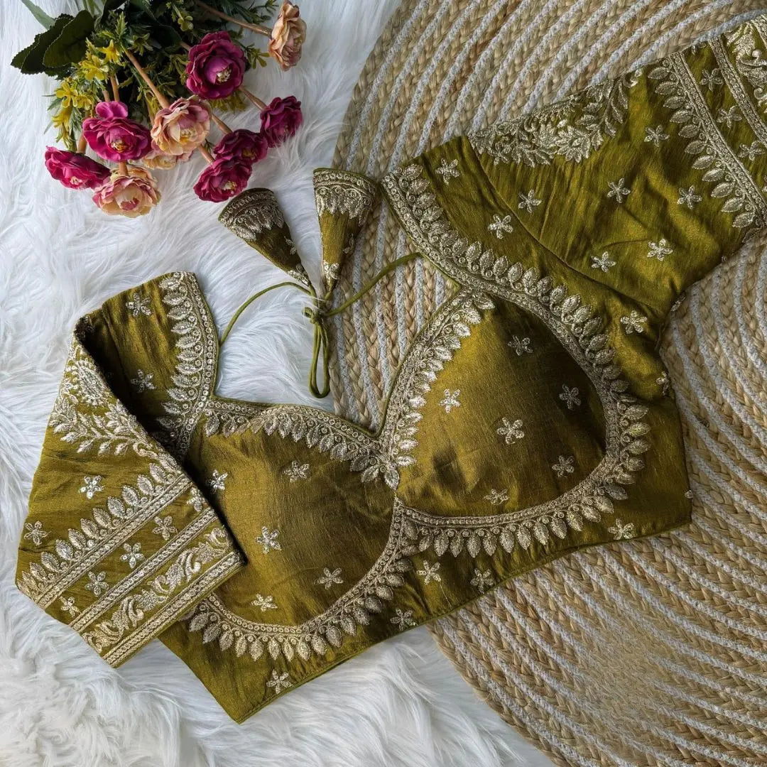 Olive green embroidered silk blouse with intricate gold zari floral patterns and tie-back detail, displayed on a white fur background with a bunch of pink and peach flowers in the corner
