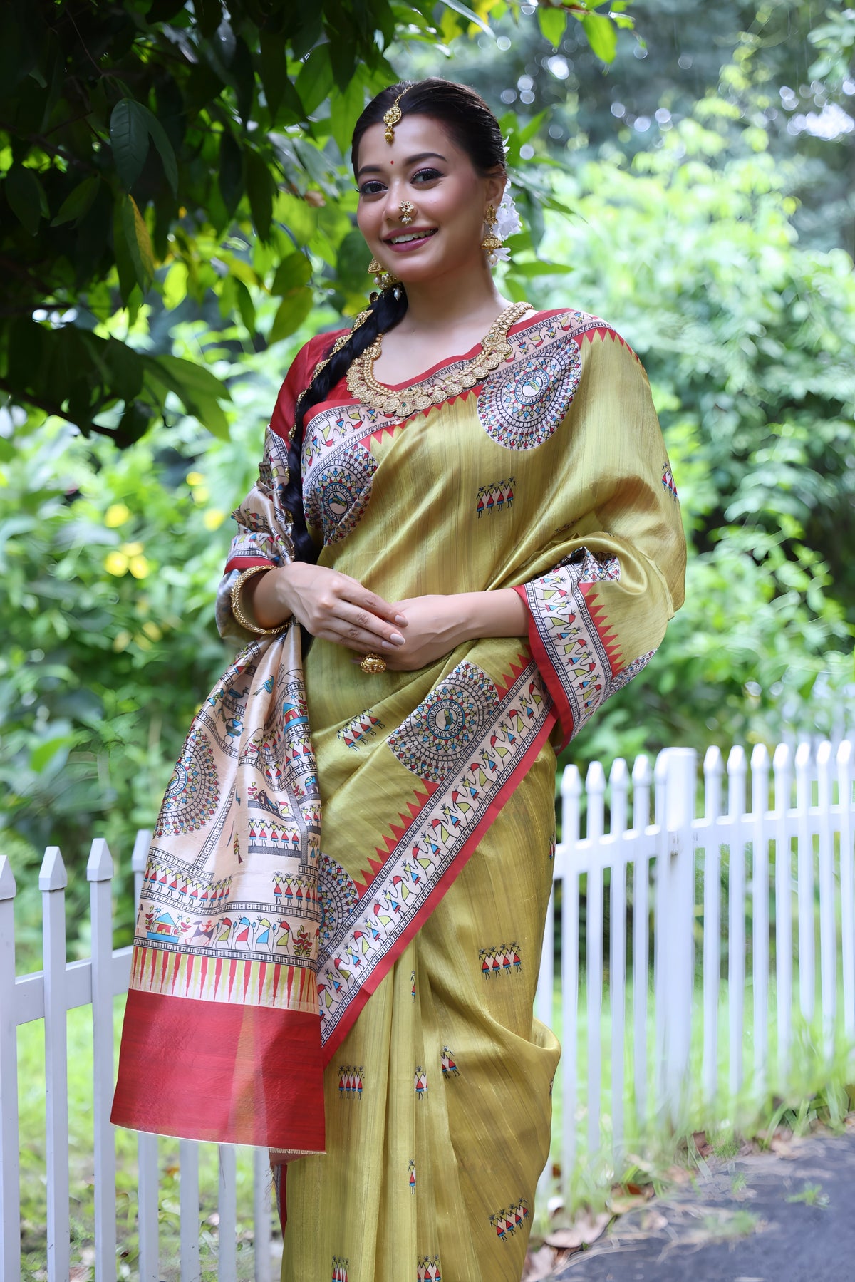 A woman models a striking Olive Green Soft Tussar Silk Saree with a red accent border. The pallu features a wide, elaborate panel with intricate White Madhubani-style folk art prints and a solid red hem. She wears a red blouse and a heavy gold choker necklace, posing outdoors.