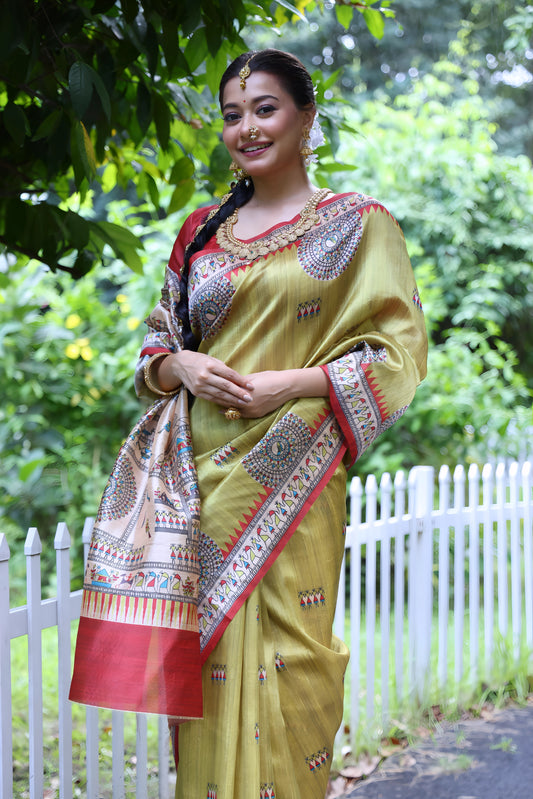 A woman models a striking Olive Green Soft Tussar Silk Saree with a red accent border. The pallu features a wide, elaborate panel with intricate White Madhubani-style folk art prints and a solid red hem. She wears a red blouse and a heavy gold choker necklace, posing outdoors.