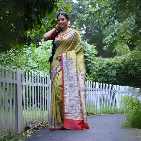 A woman models a striking Olive Green Soft Tussar Silk Saree with a red accent border. The pallu features a wide, elaborate panel with intricate White Madhubani-style folk art prints and a solid red hem. She wears a red blouse and a heavy gold choker necklace, posing outdoors.