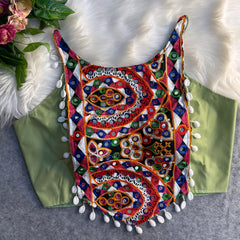 Traditional olive green blouse featuring vibrant multicolor mirror work and intricate thread embroidery in geometric and paisley patterns, accented with white shell tassels along the hem, displayed flat on a soft fur and grey background with floral and leafy decor.