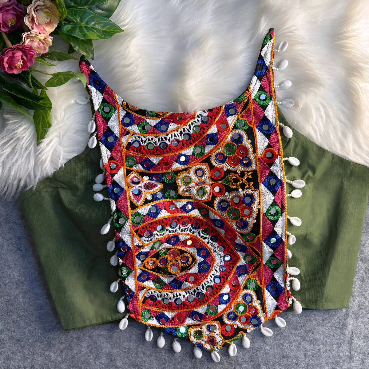 Traditional olive green blouse featuring vibrant multicolor mirror work and intricate thread embroidery in geometric and paisley patterns, accented with white shell tassels along the hem, displayed flat on a soft fur and grey background with floral and leafy decor.