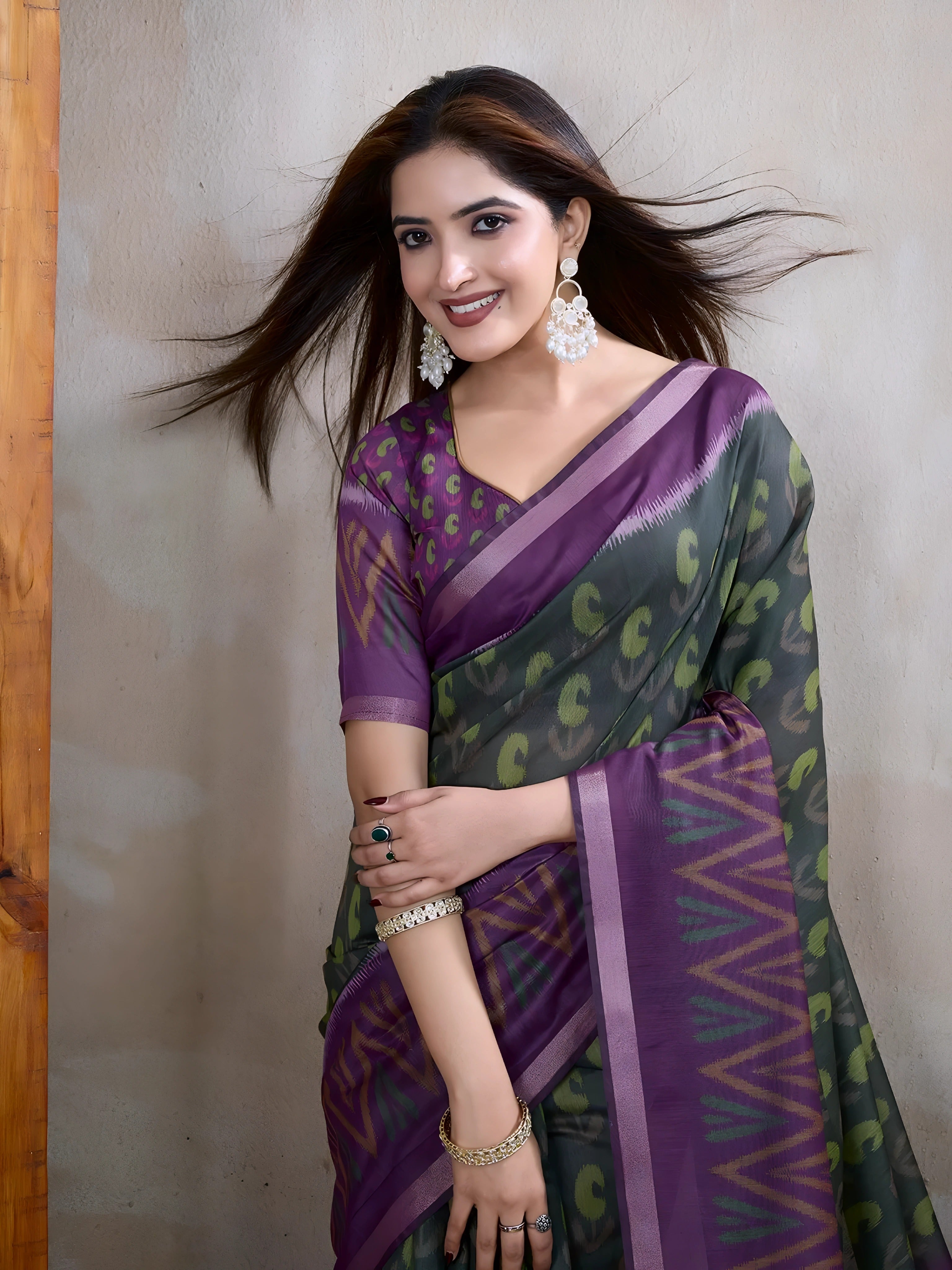  Woman wearing an olive green saree adorned with purple geometric patterns and bold abstract motifs, paired with a matching blouse and statement silver earrings. She poses elegantly in a contemporary studio setting against a neutral textured wall, with a tall woven screen and rustic wooden flooring, showcasing the saree’s vibrant pallu and intricate design.