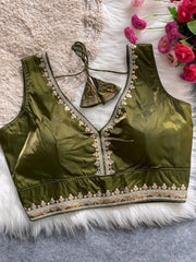 Traditional olive green silk blouse featuring intricate pastel embroidery along the neckline and hem, with a decorative tassel tie at the back, displayed flat on a soft fur and textured mat background with artificial pink flowers in the corner.