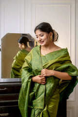 A woman models a luxurious Olive Green Raw Silk Saree. The saree features a wide border and pallu with dense, intricate tone-on-tone Kalamkari-style weaving in a matching green Zari. She wears a long-sleeve green blouse and subtle gold jewelry, with white flowers adorning her hair, posing indoors next to a dark wooden vanity.