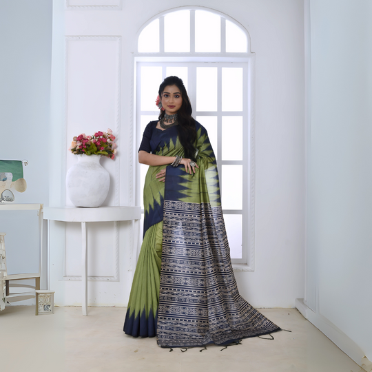 A woman models a striking Pista Green Soft Tussar Silk Saree. The saree features a bold black/navy zigzag or V-shaped pattern along the border and transitions on the body. The pallu has a wide panel of dense tribal or Ikat-style geometric print in light grey and white on a dark background. She wears a black/navy blouse and chunky oxidized silver jewelry, posing indoors by a large arched window.