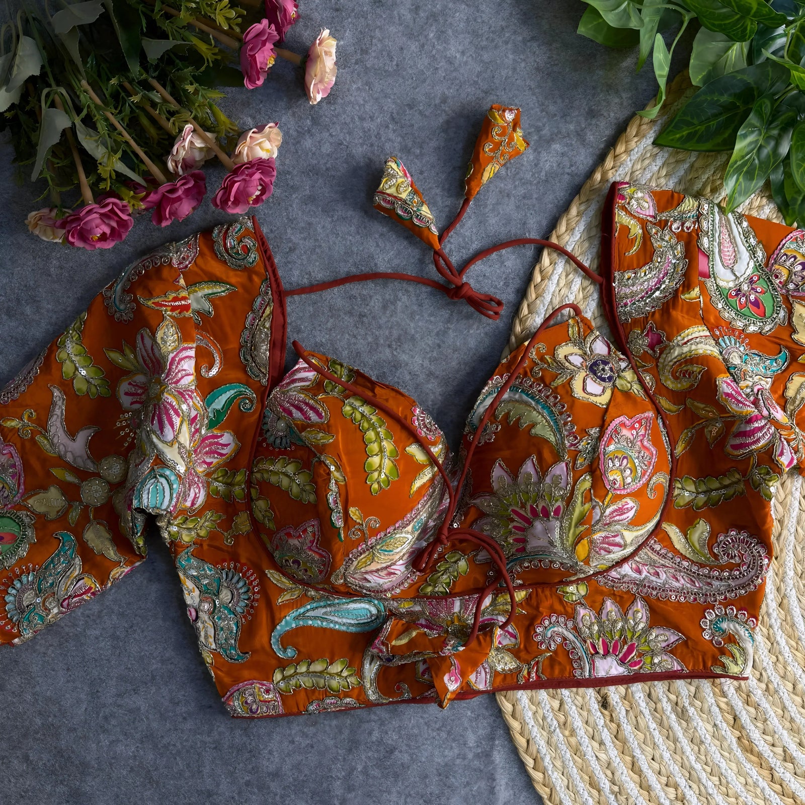  Orange padded blouse with multicolor floral and paisley embroidery, featuring sequin accents and a tie-back detail, displayed on a grey surface with artificial pink flowers and green leaves in the background.