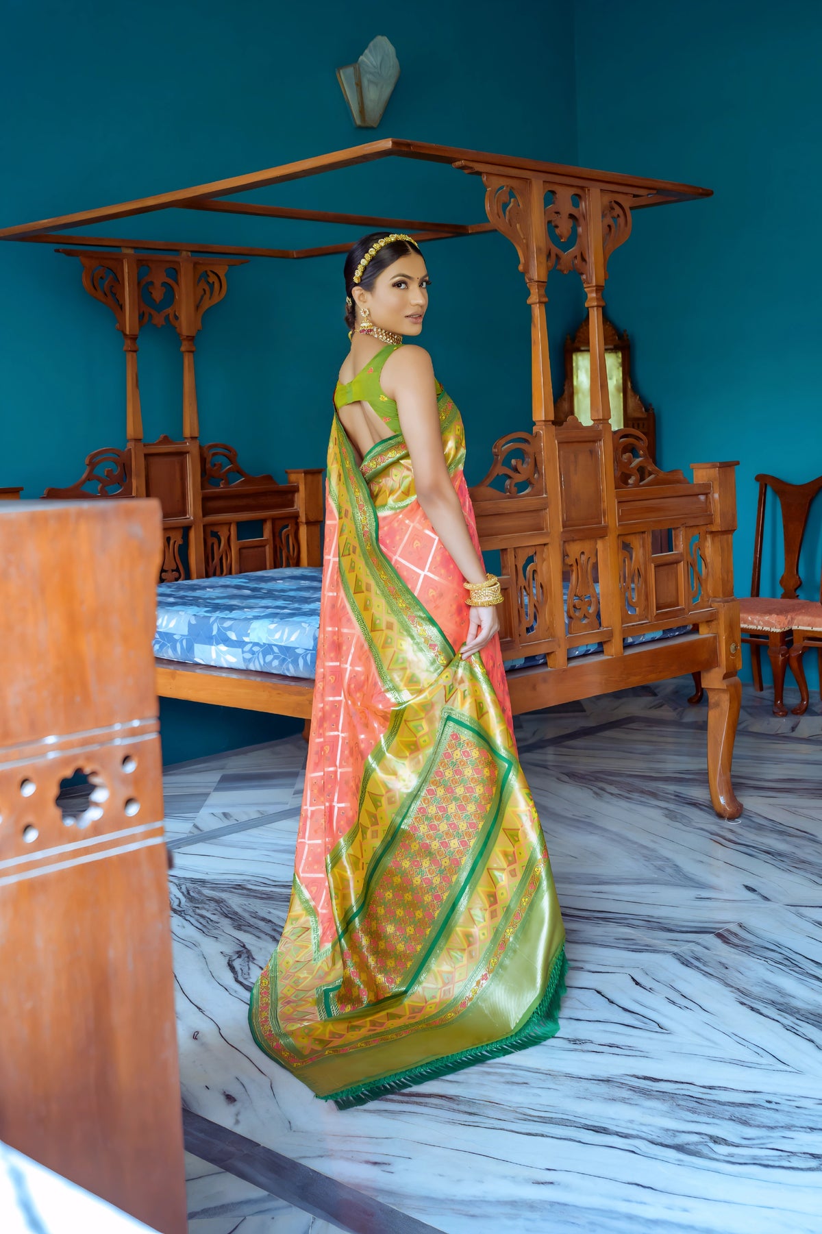 A woman models a striking Peach/Coral Banarasi Silk Saree with all-over Zari weaving. The wide pallu and border contrast dramatically in Olive Green with large geometric Zari weaving. She wears a green blouse, traditional gold jewelry, and a metallic waist belt, posing in an ornate room.