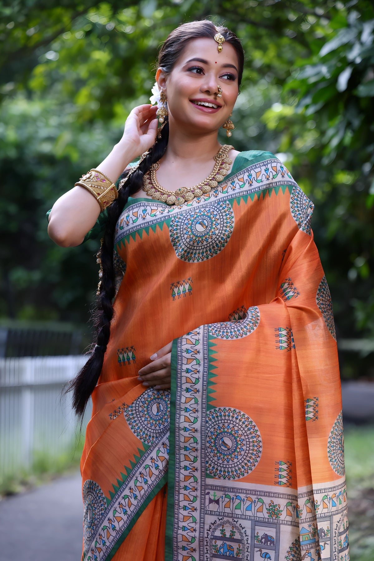 A woman models a vibrant Coral Orange Soft Tussar Silk Saree. The saree has a deep green border and an elaborate pallu featuring intricate White Madhubani-style folk art prints with blue and green figurative motifs. She wears a green embellished blouse and a gold Kundan choker necklace.