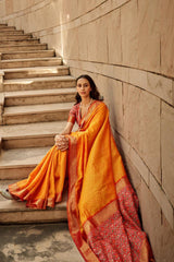 Woman in orange red saree featuring intricate zari work and designer pallu sitting gracefully on stone stairs in ethnic fashion attire