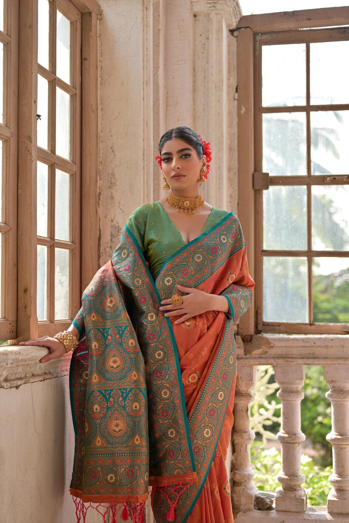 A woman models a vibrant Coral Orange Banarasi Silk Saree with all-over gold Zari weaving. The saree has a wide, ornate Rama Blue/Teal border featuring traditional Zari weaving. She wears a light green blouse and heavy gold Kundan jewelry, posing by a large antique window.