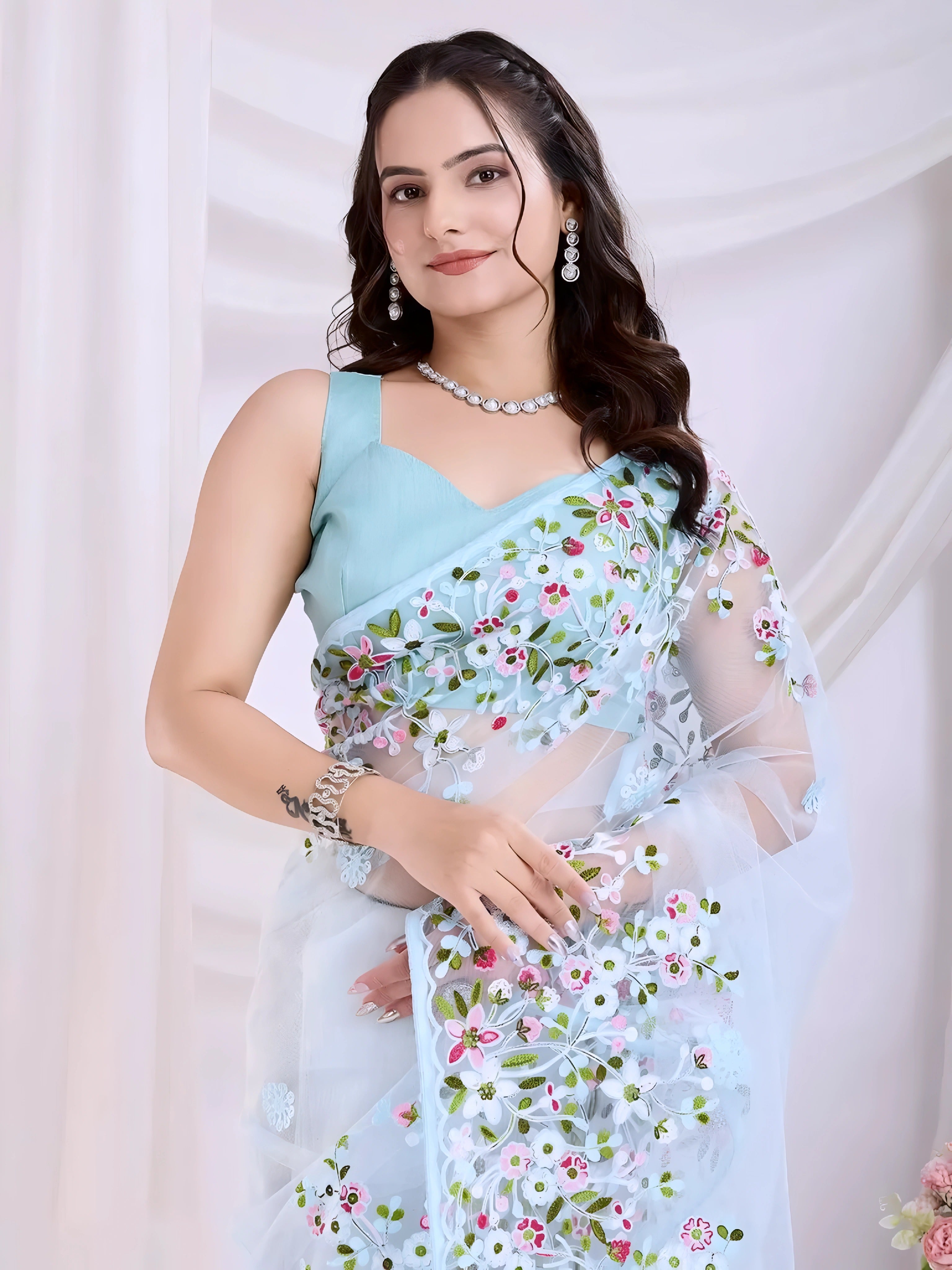 A woman models an ethereal Light Blue Soft Net Saree. The sheer fabric is densely covered in delicate all-over floral embroidery featuring white, pink, red, and green threadwork. She wears a matching sleeveless light blue blouse and accessorizes with sophisticated silver/diamond jewelry, posing indoors against sheer white curtains.