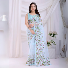 A woman models an ethereal Light Blue Soft Net Saree. The sheer fabric is densely covered in delicate all-over floral embroidery featuring white, pink, red, and green threadwork. She wears a matching sleeveless light blue blouse and accessorizes with sophisticated silver/diamond jewelry, posing indoors against sheer white curtains.
