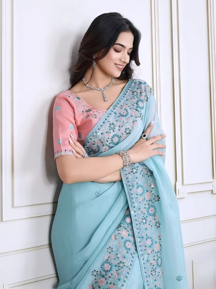  Woman wearing a pastel blue saree with intricate floral embroidery on the border, paired with a pink embroidered blouse, posiA woman models an ethereal Light Sea Blue Silk Saree. The saree features a wide, heavy border and pallu of intricate floral and vine embroidery in soft pink, teal, and silver threadwork. She wears a contrasting short-sleeve soft pink embroidered blouse and accessorizes with delicate silver/diang in a modern white interior with a wooden chair, decorative table, and floral arrangement.