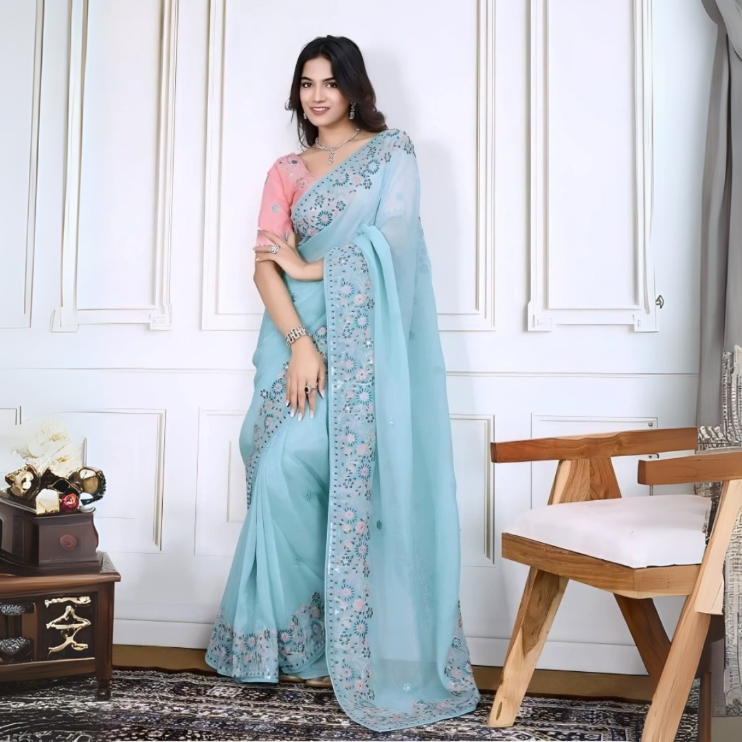 A woman models an ethereal Light Sea Blue Silk Saree. The saree features a wide, heavy border and pallu of intricate floral and vine embroidery in soft pink, teal, and silver threadwork. She wears a contrasting short-sleeve soft pink embroidered blouse and accessorizes with delicate silver/diamond jewelry, posing indoors.